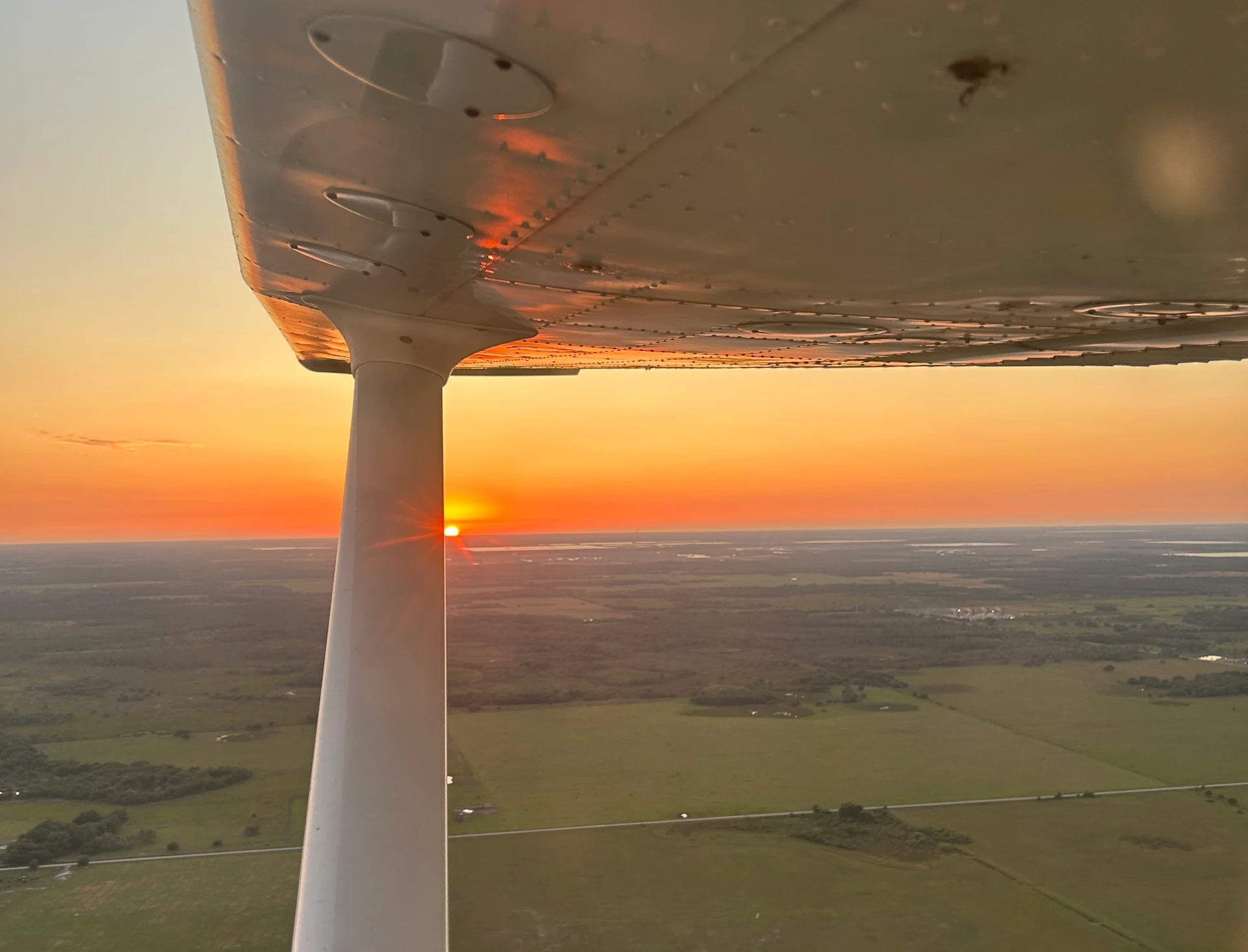Plane flying over land with sunset in the background seen from cockpit window, airplane window visible above